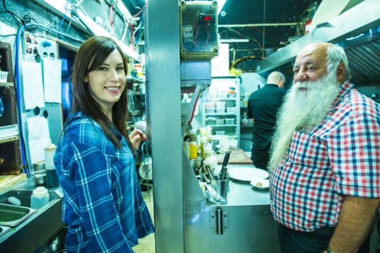 A man with a white beard (Chef Uri Jeremias) and a brunette woman (Tori Avey) in a restaurant kitchen.