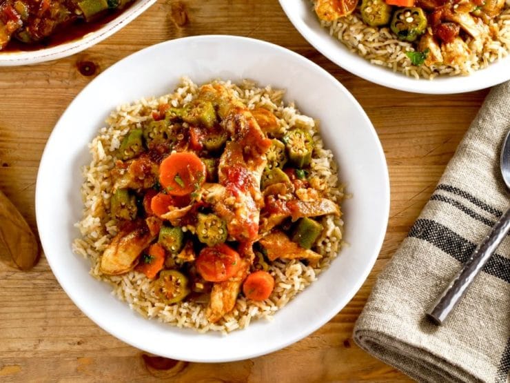 Close up overhead shot of Chicken Okra Stew on a bed of rice in a white bowl on a wooden background, two bowls of stew and cloth napkin with fork in background.