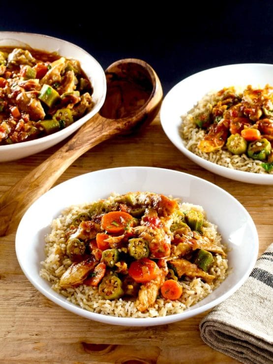 Chicken Okra Stew on a bed of rice in a white bowl on a wooden background, two bowls of stew and wooden spoon in background.