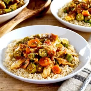 Close up of Chicken Okra Stew on a bed of rice in a white bowl on a wooden background, two bowls of stew and wooden spoon in background.