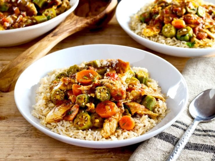 Close up of Chicken Okra Stew on a bed of rice in a white bowl on a wooden background, two bowls of stew and wooden spoon in background.
