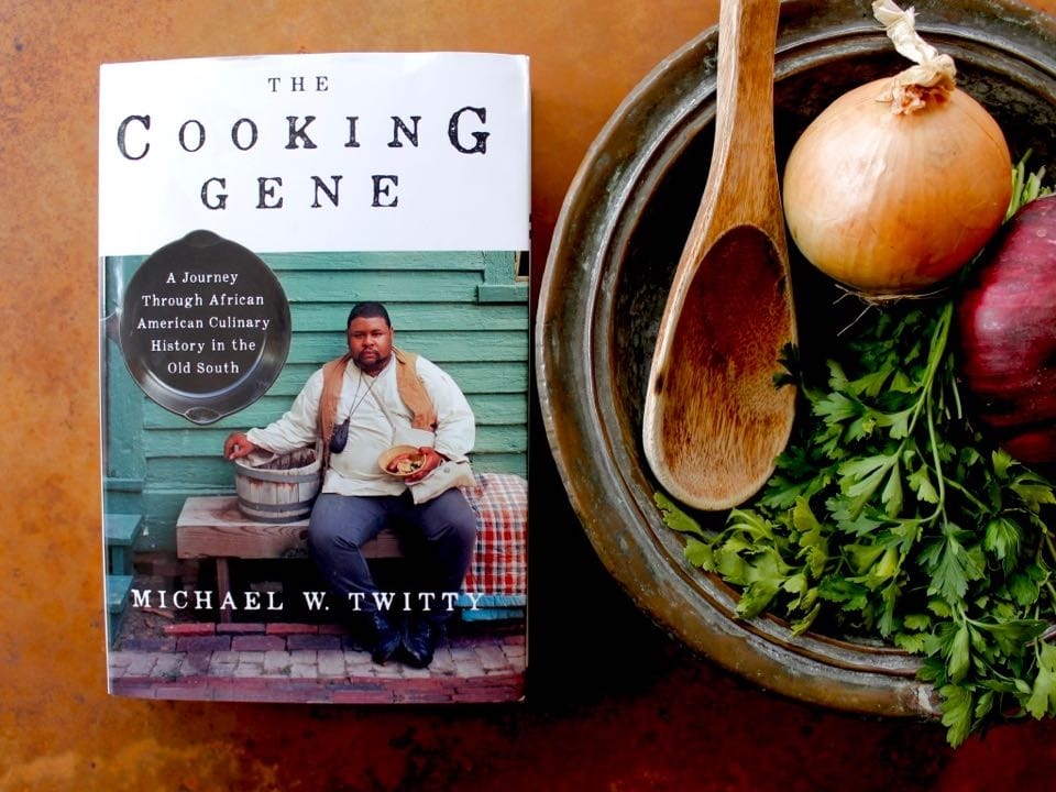 A copy of the book The Cooking Gene by Michael Twitty sits next to to a bowl filled with cilantro, a red onion, a brown onion, and a wooden spoon.