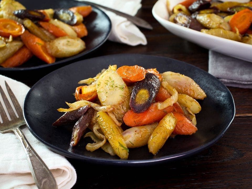 A pile of roasted carrots and fennel on a black plate next to a fork sitting on a white napkin.