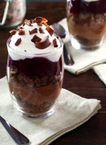 Dark chocolate cherry cheesecake parfait in a glass dessert dish on top of a white napkin next to a spoon on a wooden table.