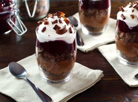 Dark chocolate cherry cheesecake parfait in a glass dessert dish on top of a white napkin next to a spoon on a wooden table.