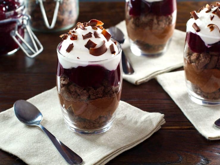 Dark chocolate cherry cheesecake parfait in a glass dessert dish on top of a white napkin next to a spoon on a wooden table.