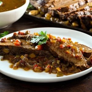 Two slices of West African Brisket on sauce topped with herbs on plate with bowl of sauce, brisket in background.