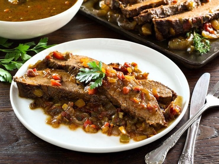 Overhead side angle shot of two slices of West African Brisket on sauce topped with herbs on plate with bowl of sauce, brisket in background.