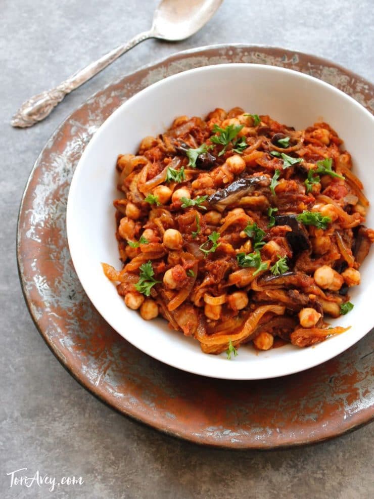 Mnazaleh - Receta con garbanzos, berenjenas y cebollas en bol blanco en bandeja de metal con cuchara.