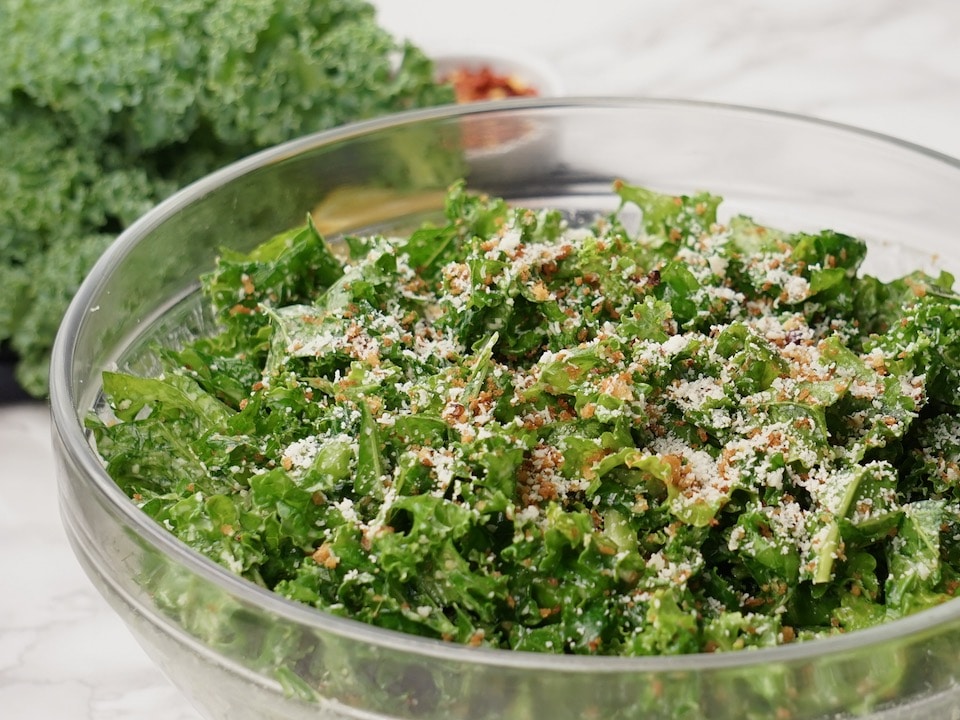 Glass bowl filled with kale Caesar salad with panko, Parmesan and red chili pepper flakes, kale leaves in background.