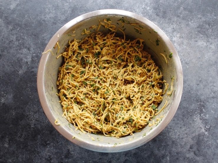 Shredded potato pancake batter mixed with tahini, herbs and spices in a large mixing bowl on concrete background.