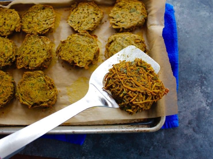 Spatula with fully cooked potato pancake over parchment lined baking sheet full of potato pancakes on concrete background with blue towel.