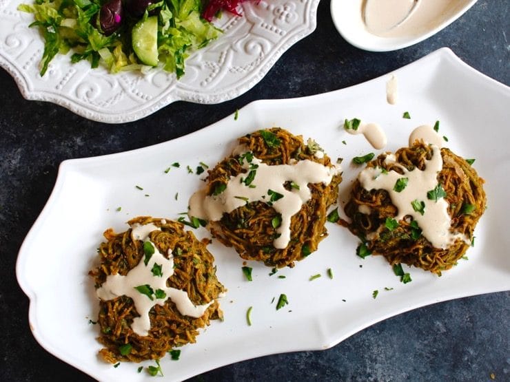 Crispy baked potato tahini pancake patties topped with tahini and parsley on white plate with bowl of tahini, spoon, and cabbage salad in background, on grey concrete counter.