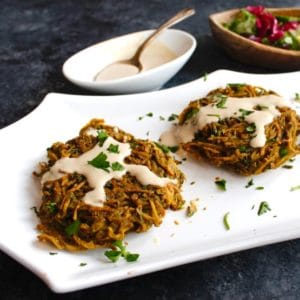 Crispy baked potato tahini pancake patties topped with tahini and parsley on white plate with bowl of tahini, spoon, and cabbage salad in background, on grey concrete counter.