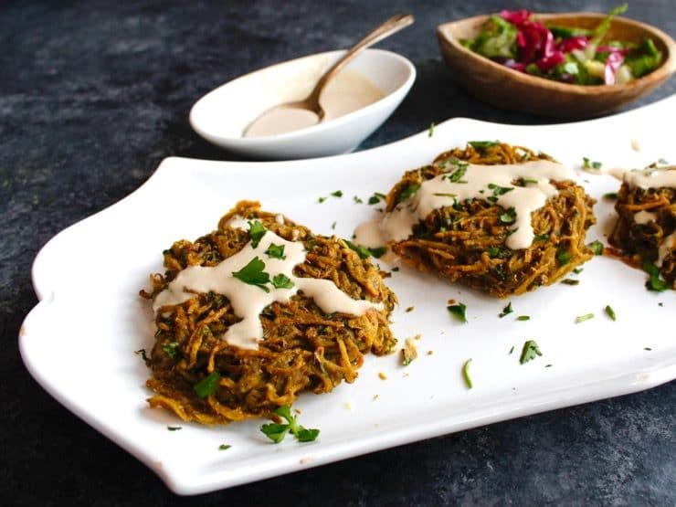 Crispy baked potato tahini pancake patties topped with tahini and parsley on white plate with bowl of tahini, spoon, and cabbage salad in background, on grey concrete counter.