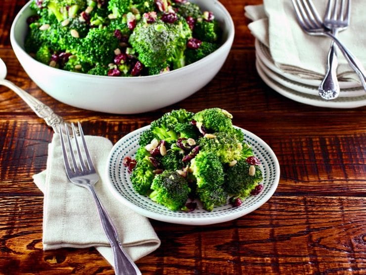 A plate of broccoli salad next to a fork and napkin on a wooden table. In the background is a large bowl of additional broccoli salad.