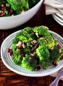 A plate of broccoli salad next to a fork and napkin on a wooden table. In the background is a large bowl of additional broccoli salad.