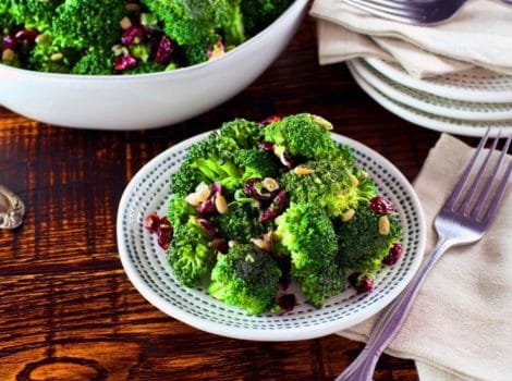A plate of broccoli salad next to a fork and napkin on a wooden table. In the background is a large bowl of additional broccoli salad.