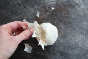 Hand removing papery skin from the outside of garlic head on grey countertop.