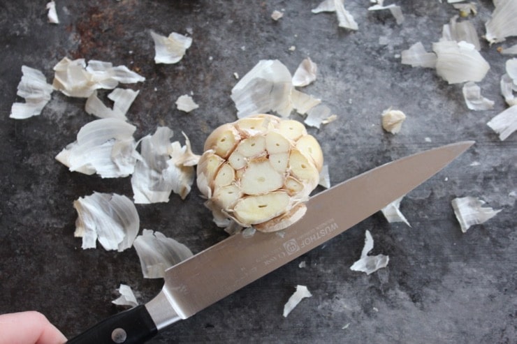 Head of garlic with exposed cloves, tips of cloves sliced off, knife and papery garlic skin on countertop in background.