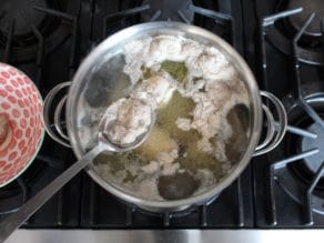 A spoon is skimming the white foam from the top of the soup stock in a large pot.