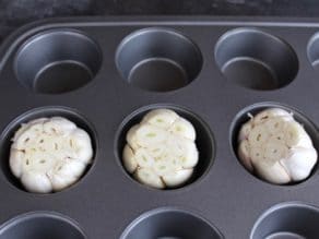 Three raw garlic heads with exposed cloves in muffin pan.