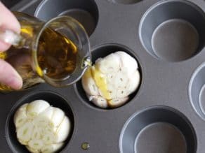 Small carafe drizzling olive oil on top of whole garlic head with exposed cloves in muffin pan.
