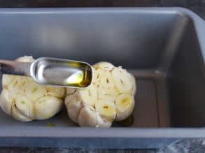 Teaspoon drizzling olive oil over two heads of raw garlic with exposed cloves in loaf pan.