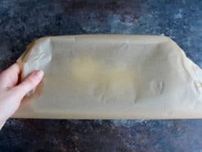Small loaf pan, hand covering with parchment on grey countertop.