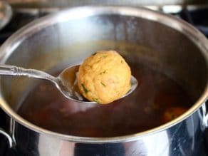 A spoon holding a matzo ball over a large pot of soup broth.