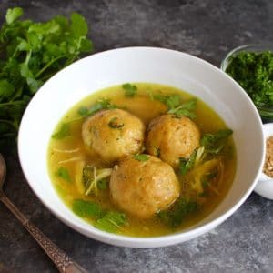 Yemenite matzo ball soup with three large matzo balls in a white bowl on a silver spoon surrounded with vegetables