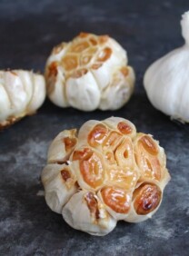 Head of roasted garlic with exposed cloves in foreground, three heads of garlic in background, one raw two roasted, on grey countertop.