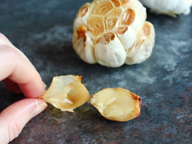 Single roasted garlic clove being squeezed from its papery casing on grey countertop, head of roasted garlic in background.