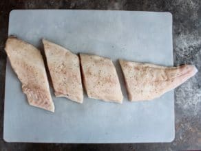 Long fresh cod fillet on cutting board, seasoned with salt and pepper, sliced into 4 pieces