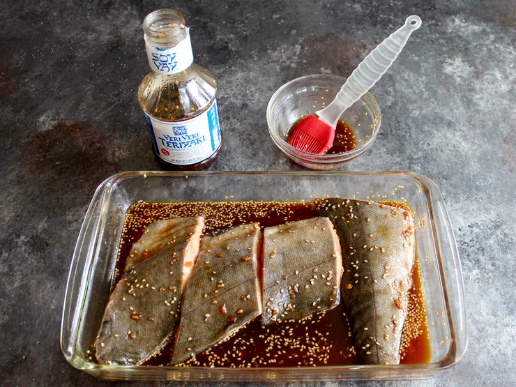 4 pieces of cod fillet in a 9x13 glass dish covered with Soy Vay Veri Veri Teriyaki Marinade and Sauce, bowl and brush with bottle of Soy Vay marinade in background.