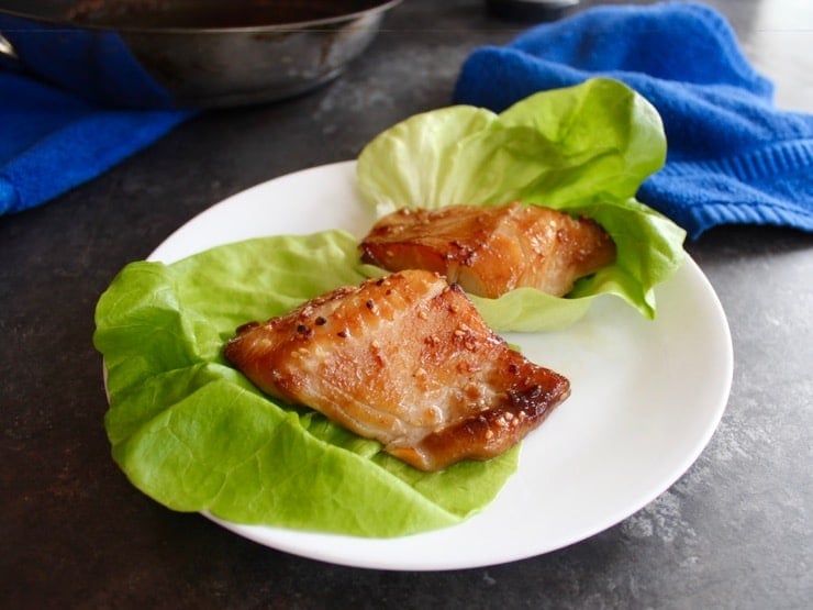 Close up on two Teriyaki Black Cod fillets, broiled and browned, on lettuce leaves on a white plate.