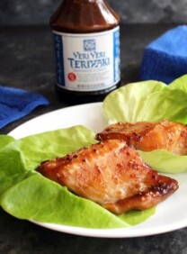 Two pieces of glazed broiled Teriyaki Black Cod in foreground, bottle of Soy Vay Veri Veri Teriyaki Marinade and Sauce in background with blue towel and skillet.