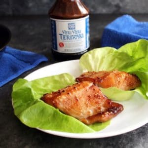 Two pieces of glazed broiled Teriyaki Black Cod in foreground, bottle of Soy Vay Veri Veri Teriyaki Marinade and Sauce in background with blue towel and skillet.