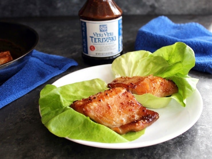 Two pieces of glazed broiled Teriyaki Black Cod in foreground, bottle of Soy Vay Veri Veri Teriyaki Marinade and Sauce in background with blue towel and skillet.