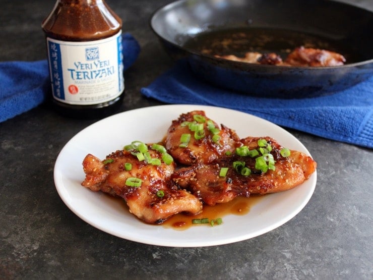 Spicy Teriyaki Broiled Chicken Thighs on white plate topped with scallions, skillet with chicken on blue towel in background with bottle of Soy Vay Veri Veri Teriyaki Marinade.