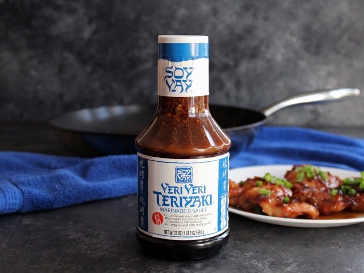 Bottle of Soy Vay Veri Veri Teriyaki Marinade on countertop with plate of Spicy Teriyaki Broiled Chicken Thighs topped with scallions in background, and skillet on blue towel.