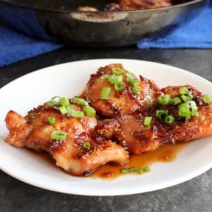 Spicy Teriyaki Broiled Chicken Thighs on white plate topped with scallions, skillet with chicken on blue towel in background.