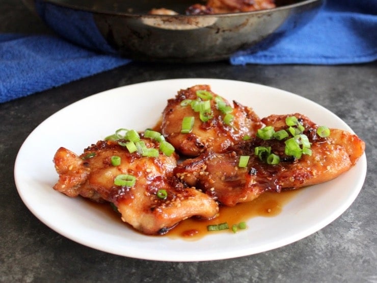 Spicy Teriyaki Broiled Chicken Thighs on white plate topped with scallions, skillet with chicken on blue towel in background.