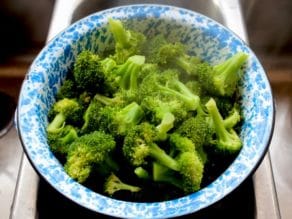 Steamed broccoli in blue speckled colander in stainless steel sink.