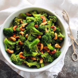 Overhead view of Citrus Broccoli with Cashews in white bowl with antique tongs on grey countertop with white linen.