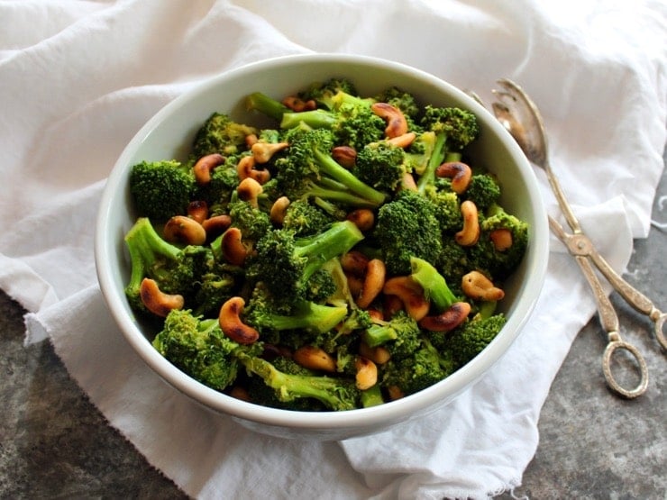 Front view of Citrus Broccoli with Cashews in white bowl with antique tongs on grey countertop with white linen.