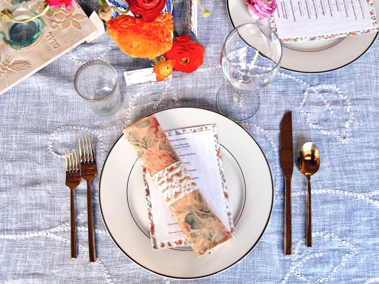 Overhead shot of table setting for Passover Seder - matzo napkin ring and matzo-backed menu on plate with cloth napkin, gold utensils, wine glass and flowers.