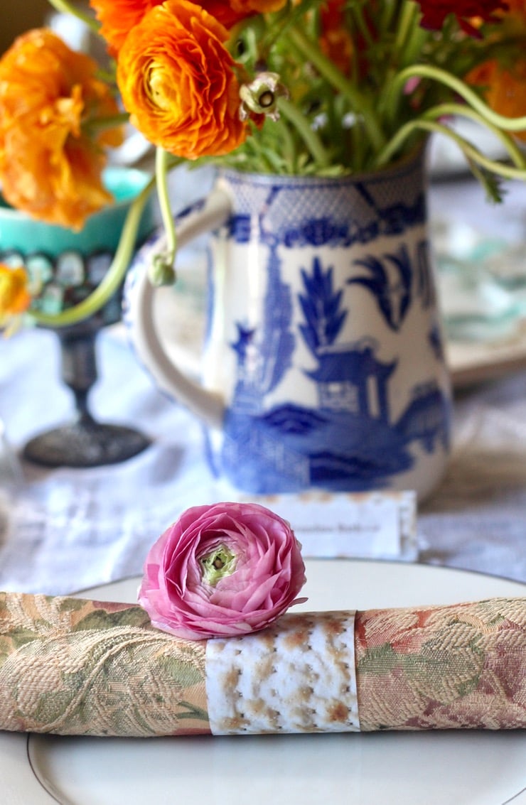 A pink ranunculus sits at the edge of a decorated Passover seder table. Behind the ranunculus is a blue and white vase and an Elijah cup.