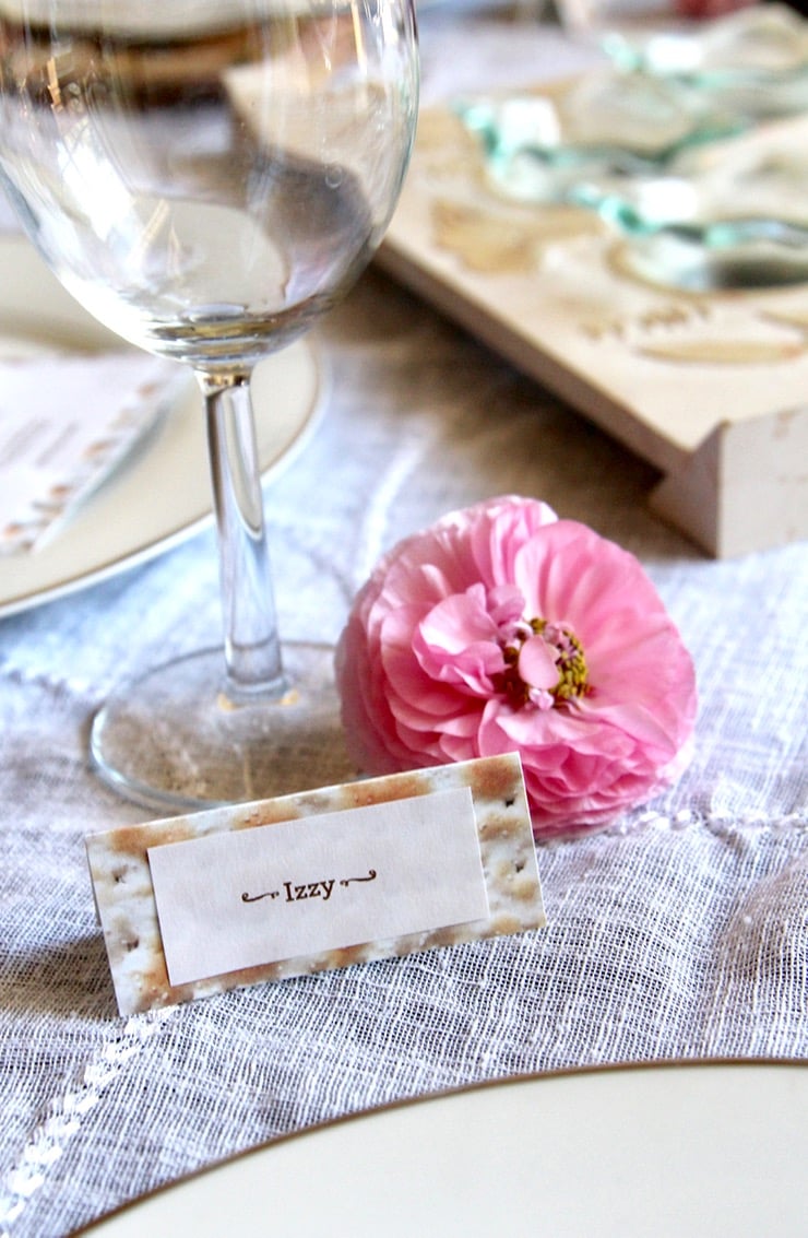 Matzo paper name tag that says "Izzy" sits in front of a wine glass and next to a pink ranunculus bud.