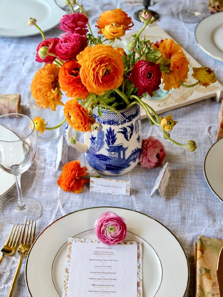 A table set with white plates a beautiful vase filled with orange and red ranunculus.